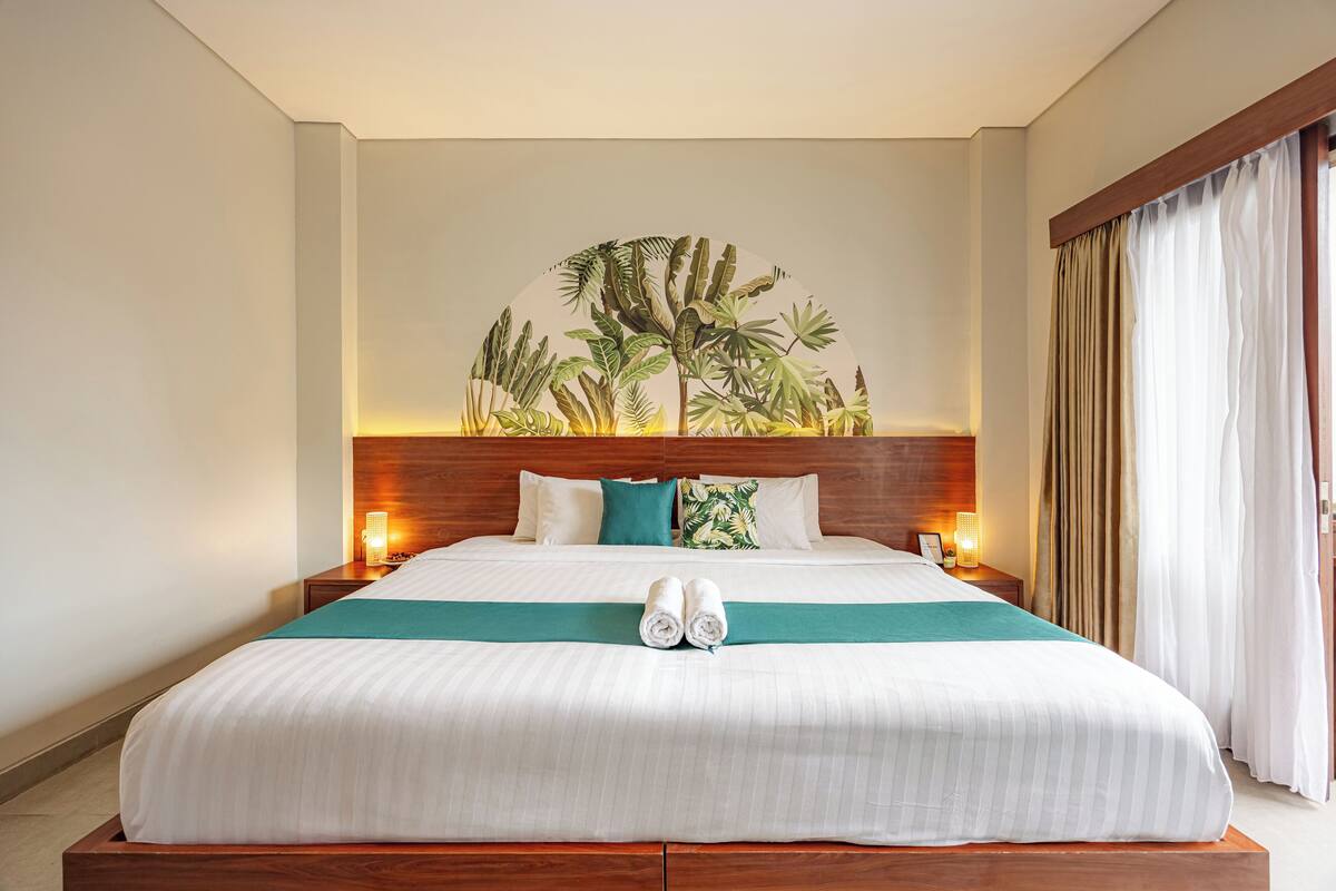 Une chambre d’hôtel avec un grand lit, une tête de lit ornée d’une fresque aux motifs tropicaux, deux lampes de chevet et une vue sur la plage par la fenêtre.