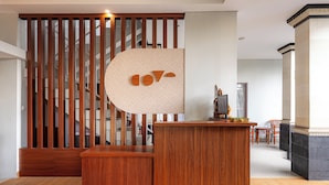 A wooden reception desk with a modern design, a staircase with wooden railings, and a view of a dining area with chairs and a table.