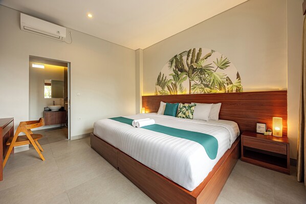 A modern hotel room with a large bed, wooden headboard, a small bedside table with a lamp, a desk with a chair, and a bathroom visible through an open door. - Cove Puspa Canggu (Tibubeneng)