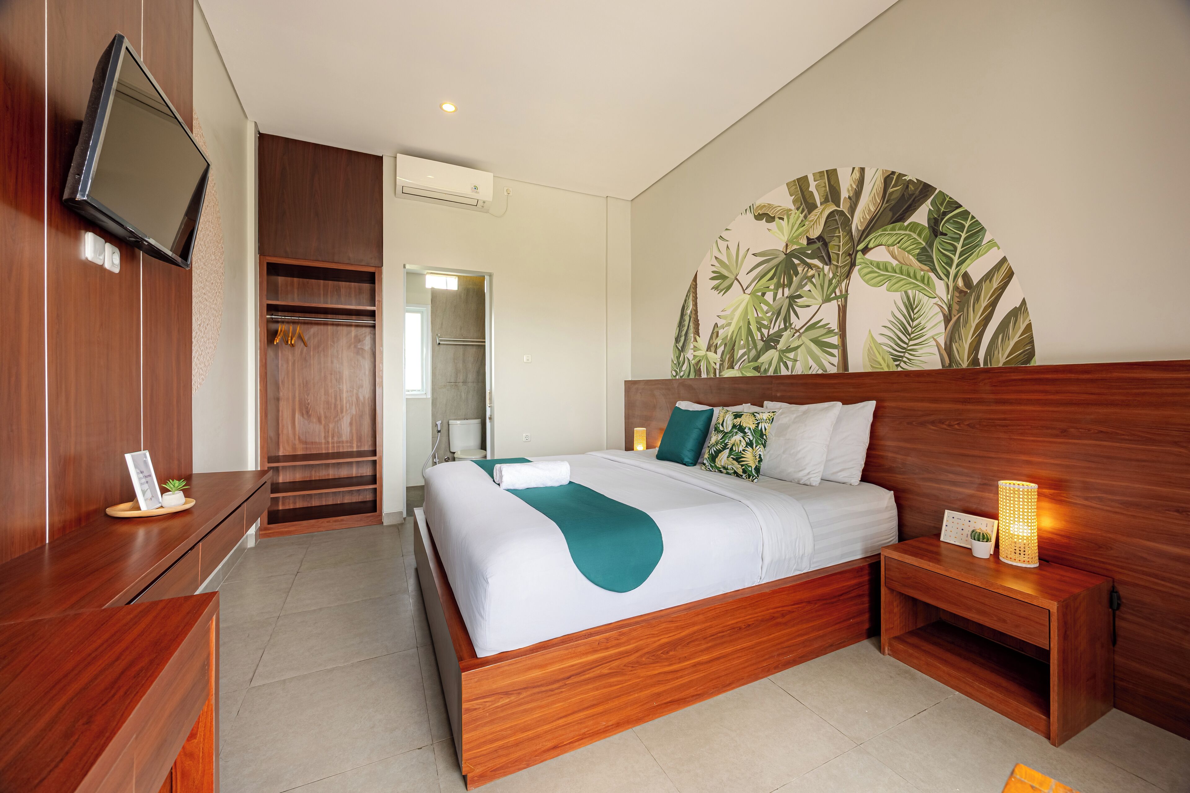 A modern hotel room with a wooden bed, a flat-screen TV, a large tropical leaf mural, and a wooden nightstand with a lamp.