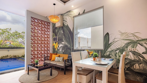 A modern living room with a glass table, wicker chairs, a sofa with cushions, a decorative wall panel, and a view of a swimming pool.