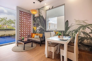 A modern living room with a glass table, wicker chairs, a sofa with cushions, a decorative wall panel, and a view of a swimming pool. - Cove Puspa Canggu (Tibubeneng)