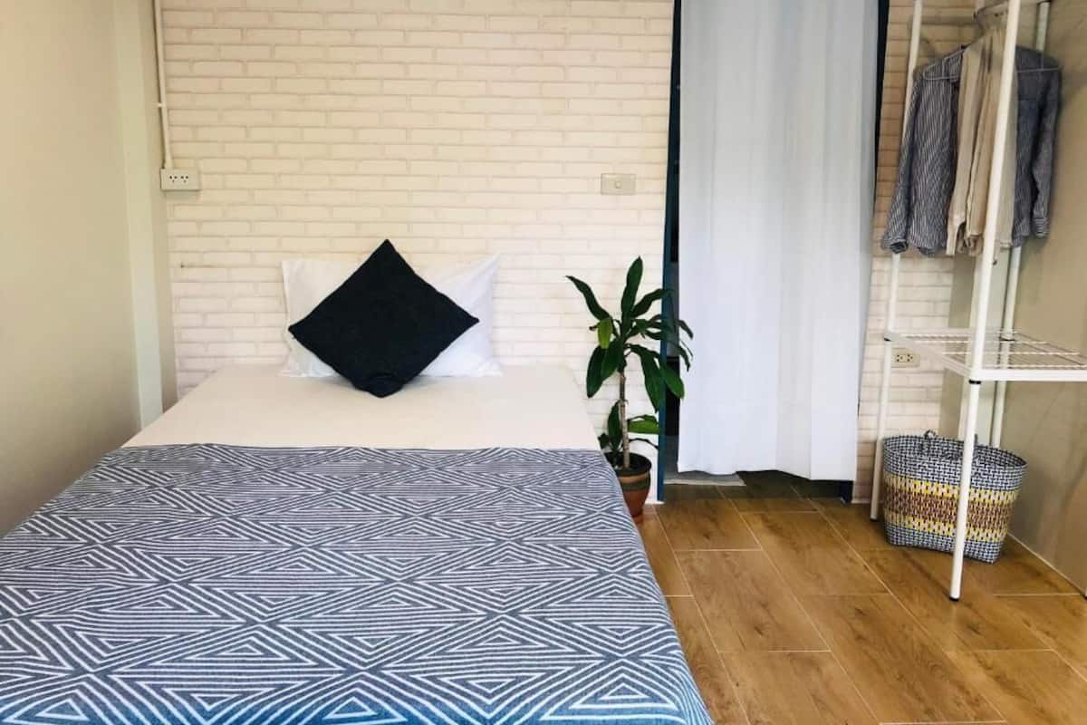 Ein Schlafzimmer mit einem Bett, einer Topfpflanze, einer weißen Ziegelwand, einem Kleiderständer und einem Korb.