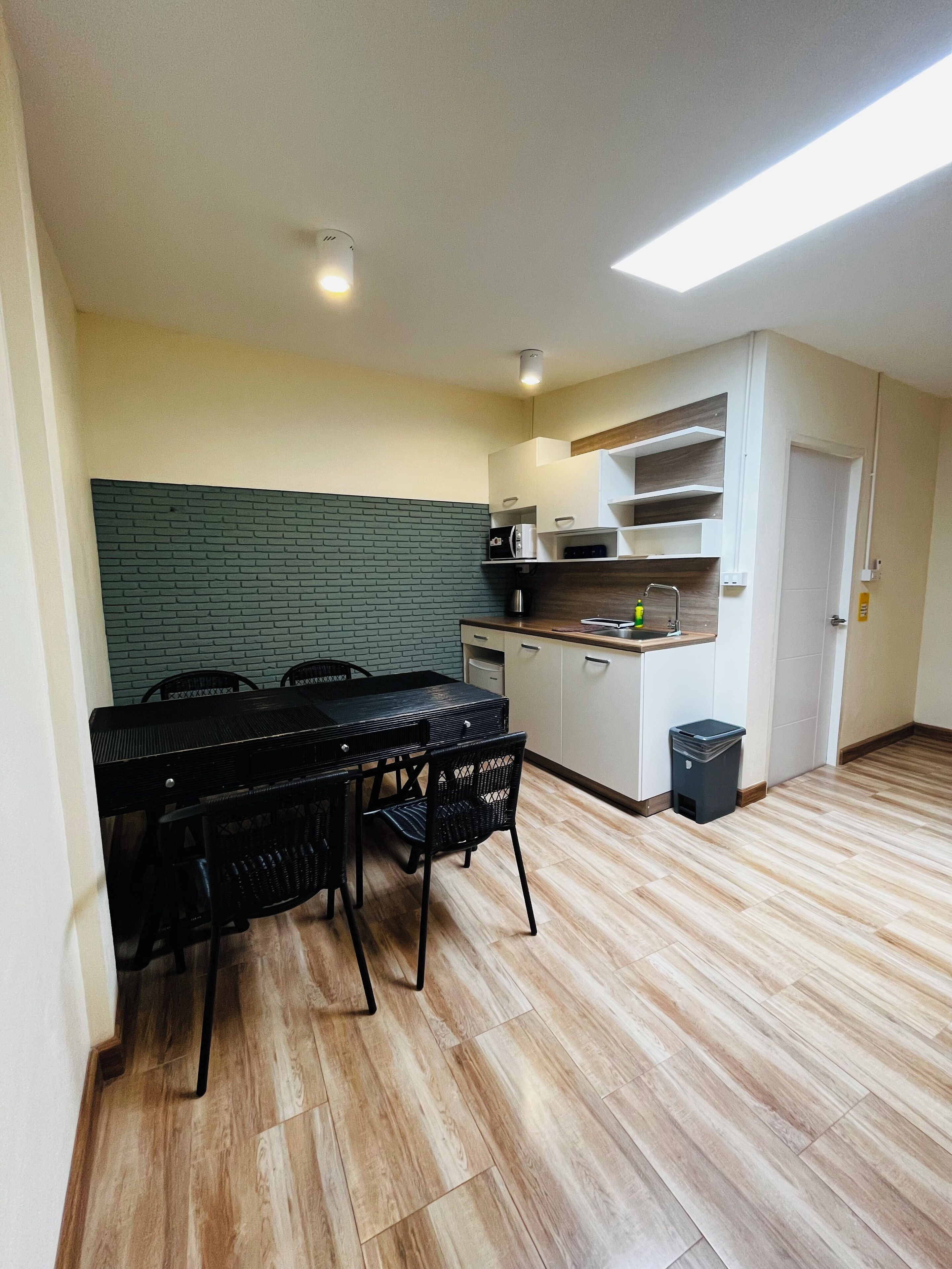 A compact kitchen with a dining area, featuring a black table and chairs, a coffee machine, and a trash bin.