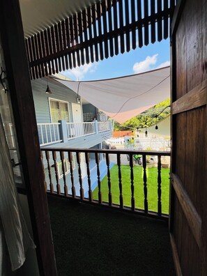 Een balkon met houten spijlen, een witte hek en uitzicht op een huis met een blauwe gevel en een witte deur.