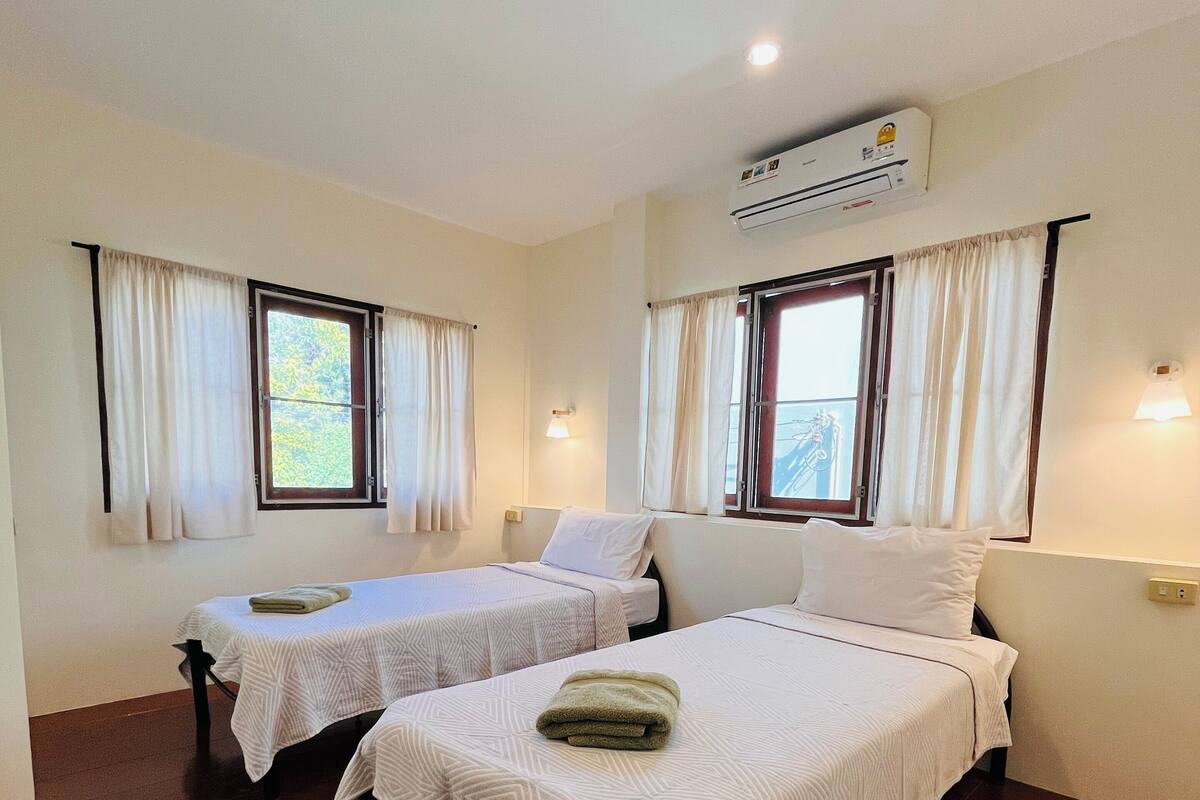 Ein Zimmer mit zwei Betten, jeweils mit weißer Bettdecke und grünem Handtuch, Holzboden und einem mit Vorhängen versehenen Fenster.