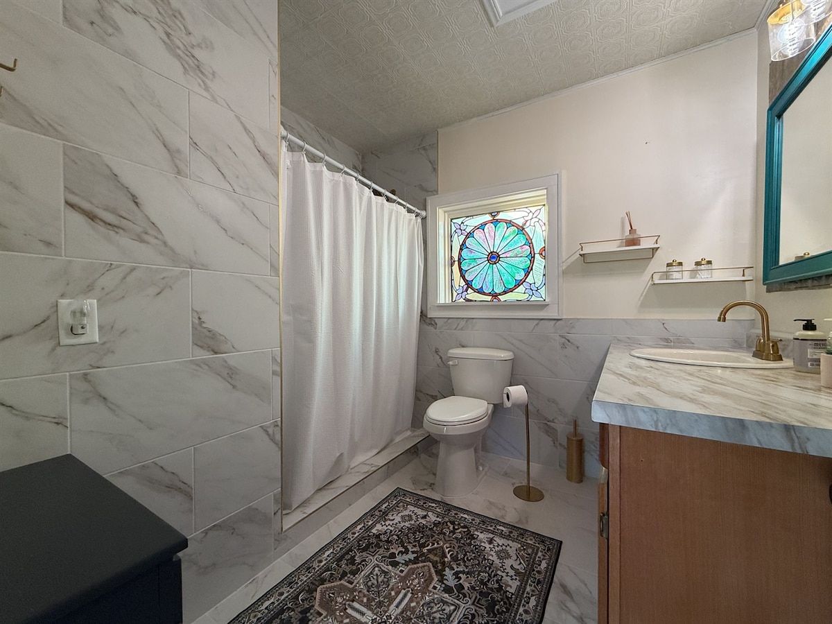 A bathroom with a toilet, a sink, a shower curtain, a patterned rug, and a stained glass window.