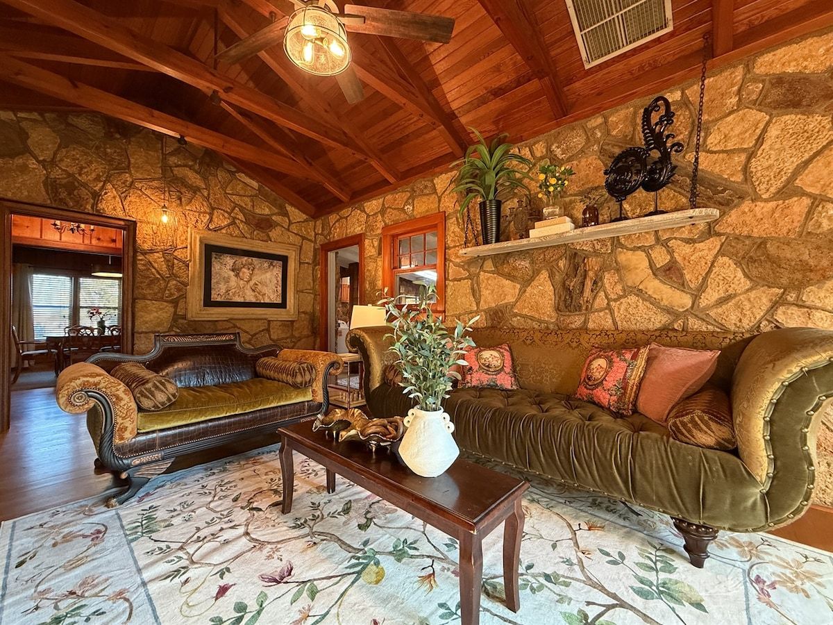 A cozy living room with a stone wall, wooden ceiling beams, a fireplace, and a large window.