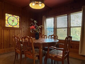 Een houten eetkamer met een ronde tafel en stoelen, een glasraam in glas-in-lood en een vaas met bloemen.