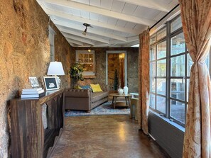 A cozy room with a stone wall, a wooden console table, a sofa, a small table with a lamp, and a large window with curtains. - La Dolce Vita - Rustic Italian-Inspired Retreat (Abilene)