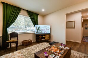 A living room with a television, a coffee table with magazines, a chair, and a window with curtains. - Red Bud Run - Stylish Abilene Retreat (Abilene)
