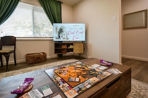 A living room with a board game, a television displaying 'Welcome Home,' and a window with curtains. - Red Bud Run - Stylish Abilene Retreat (Abilene)