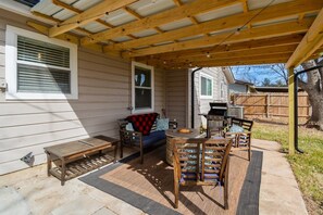 Un patio couvert avec un banc en bois, une table ronde entourée de chaises et un barbecue.