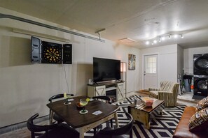 A living room with a flat-screen TV, a dartboard, a dining table with chairs, a patterned rug, a sofa, and a washing machine. - Red Bud Run - Stylish Abilene Retreat (Abilene)