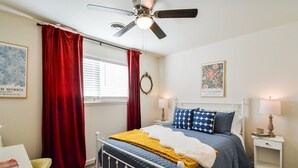 Quarto com cama, ventilador de teto, janela com cortinas, mesa de cabeceira e decoração na parede.