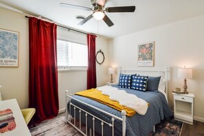 Une chambre à coucher comprenant un lit, un ventilateur de plafond, une fenêtre avec des rideaux, une table de chevet et des œuvres d’art murales.