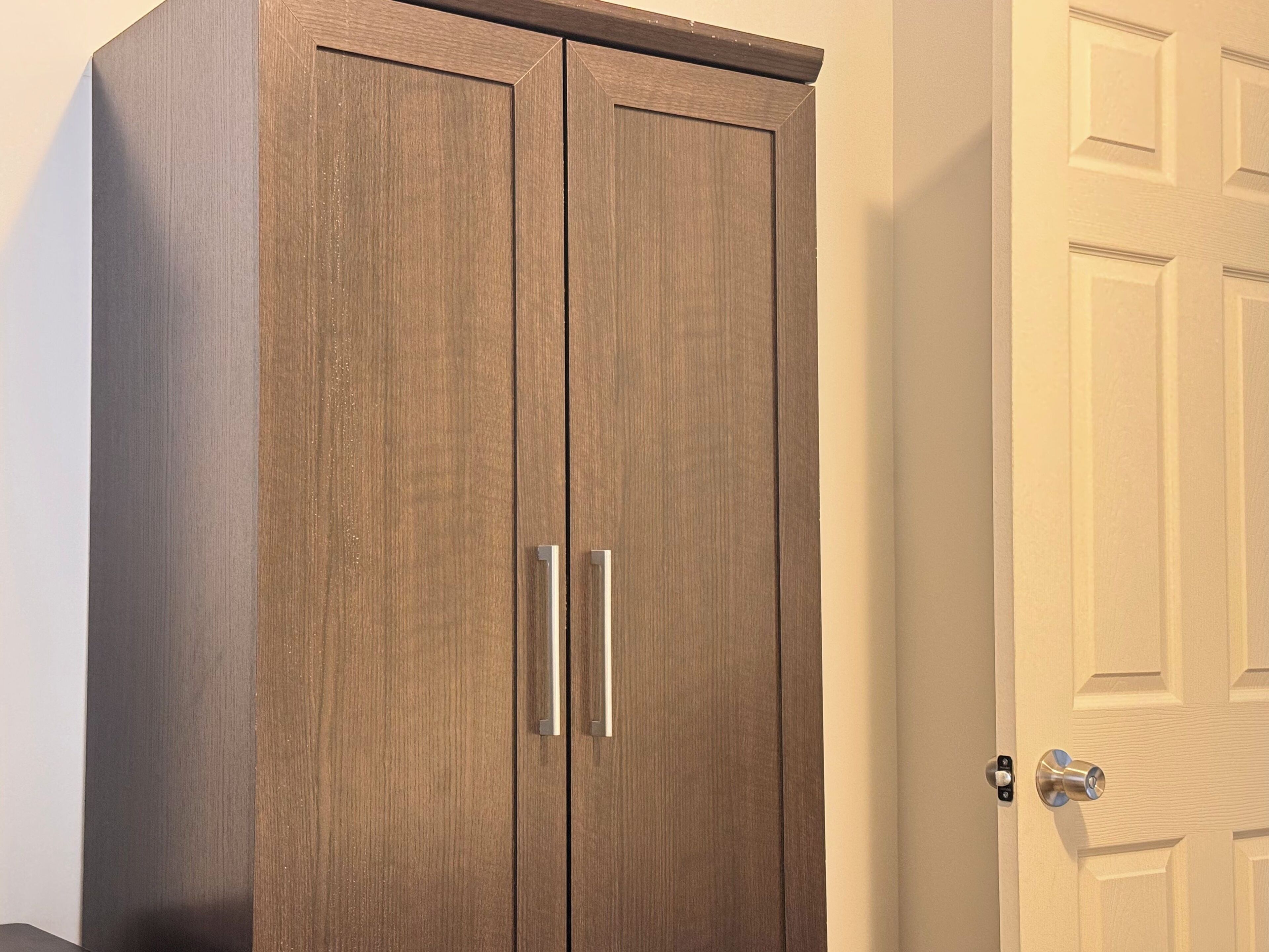 Une armoire en bois à deux portes, dotée de poignées argentées, située à côté d’une porte blanche également munie d’une poignée argentée.