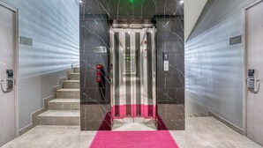 Un ascensor moderno con pantalla digital, paredes de mármol y una alfombra rosa que conduce a una escalera.