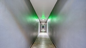 Un pasillo largo y estrecho con una señal de salida verde al final, flanqueado por paredes lisas y un suelo de baldosas.