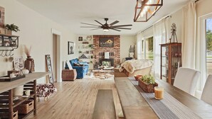A spacious living room with a fireplace, a ceiling fan, and a large window with curtains. - Wellness at the Ranch Retreat! 2 Bedroom 2 Bath Oasis with outdoor living space (Acampo)