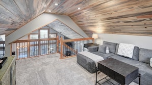 Quarto em estilo loft com teto inclinado, escada de madeira, sofá cinza e vista para o exterior.