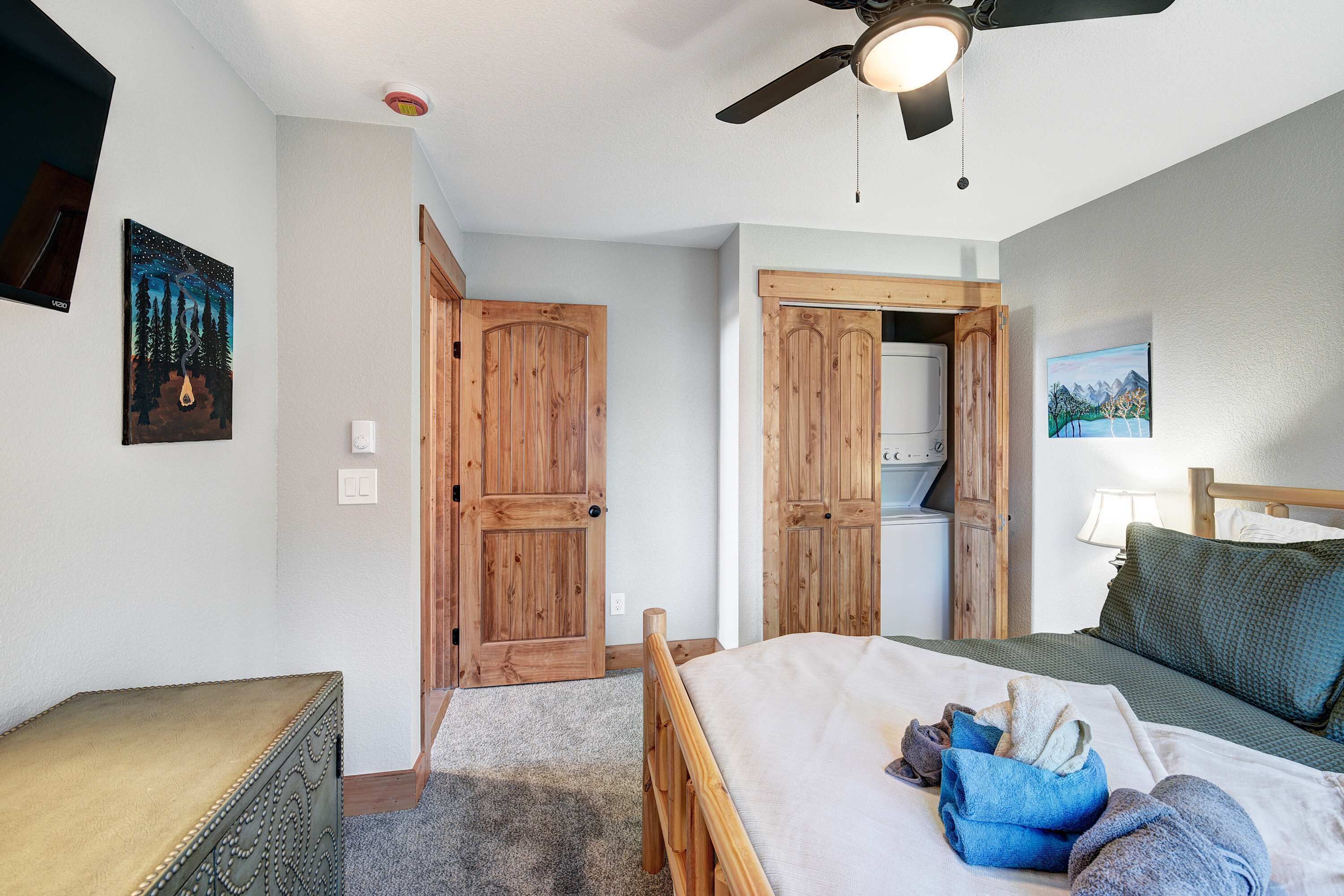 A bedroom with a bed, a wooden wardrobe, a ceiling fan, and a painting on the wall.