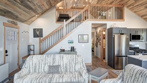 Uma sala de estar ampla com teto alto, vigas de madeira, escada, sofá, mesa de centro e área de cozinha com eletrodomésticos modernos.