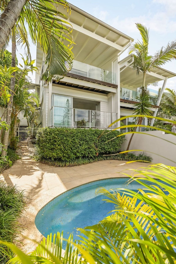 A modern two-story house with a swimming pool, surrounded by lush greenery and palm trees.