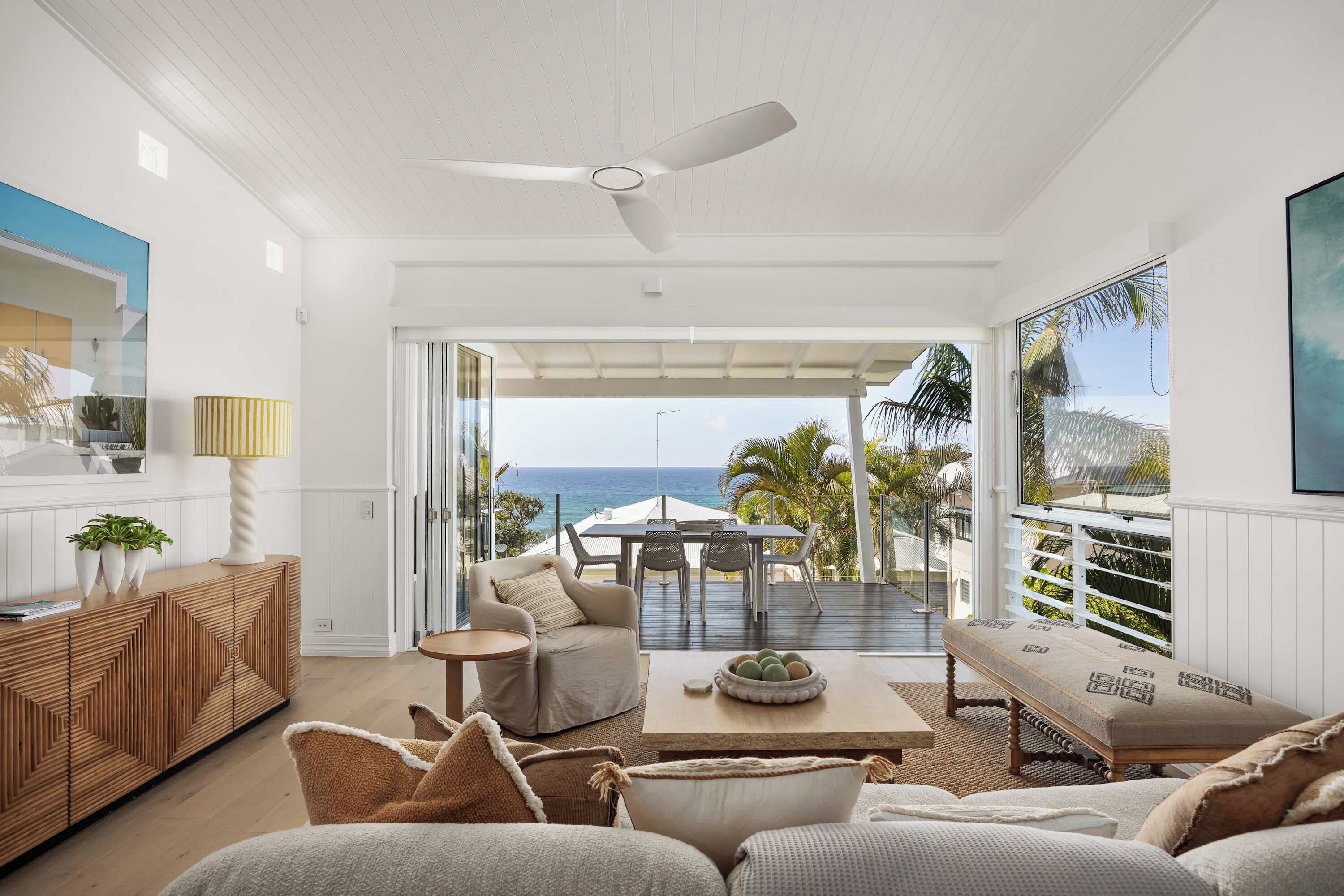 A modern living room with a view of the ocean, a ceiling fan, and a dining area with outdoor seating.