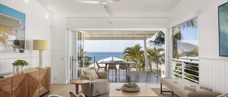 A modern living room with a view of the ocean, a ceiling fan, and a dining area with outdoor seating.