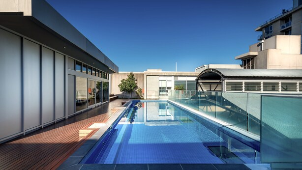 A modern rooftop pool with glass railings, wooden deck, and a clear blue sky. - Mega Style Apartments Tribeca (East Melbourne)