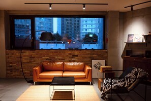 Un salon moderne avec un canapé en cuir, une table basse, une vue sur le paysage urbain à travers une grande fenêtre et un mur en briques.