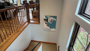 Una scala con ringhiere in legno conduce a una zona living con camino in pietra, un'area pranzo con sedie e un poster del Montana appeso al muro.