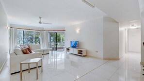 A modern living room with a white sofa, a TV on a stand, a ceiling fan, and large windows. - Ulysses 2 Beach front Apartment (Wongaling Beach)
