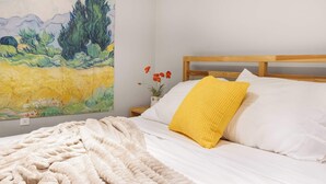 Un dormitorio con una cama, un almohadón amarillo, una manta blanca y un cuadro en la pared.