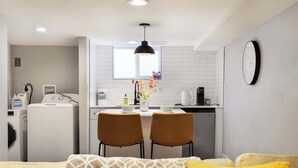 Un espacio habitable compacto con kitchenette, una zona de comedor con dos sillas, un sofá con cojines estampados y un reloj montado en la pared.