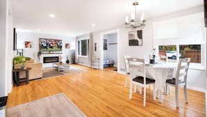 A spacious living room with a fireplace, a dining area with a white table and chairs, and a large TV mounted on the wall. - Modern 2BR Home in Ravenna, Near UW, Green Lake (Seattle)