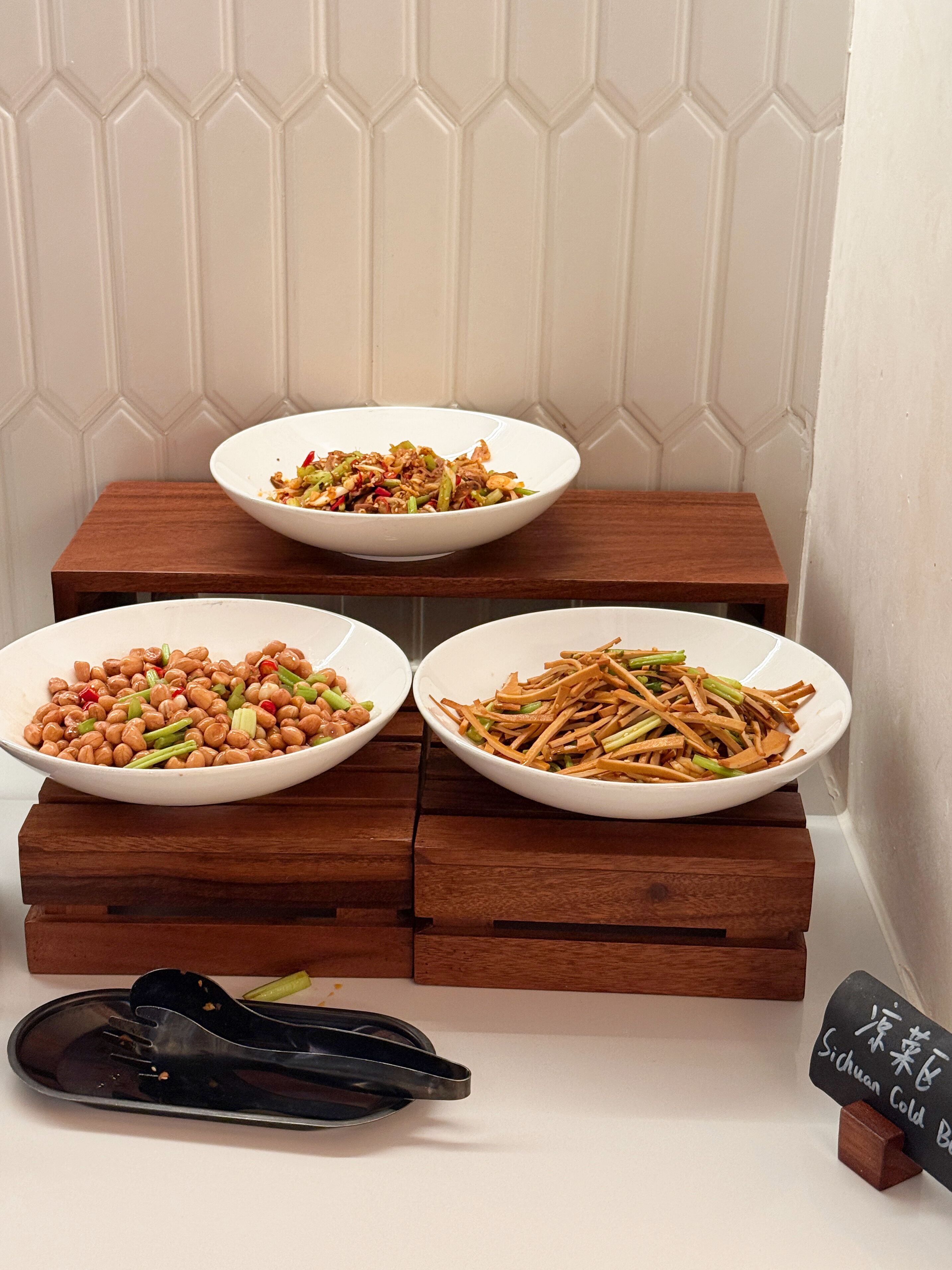 Drei weiße Schüsseln mit Essen auf Holztischen, eine schwarze Ablage mit Stäbchen und ein Schild mit chinesischen Schriftzeichen.