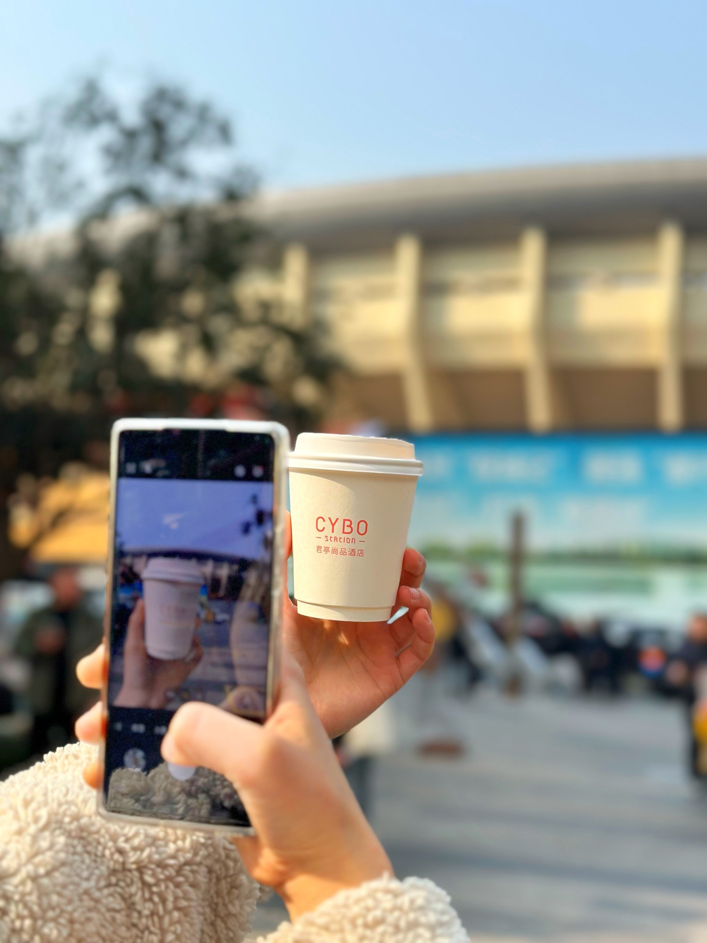 A person holding a coffee cup with 'CYBO' branding, taking a photo with a smartphone.
