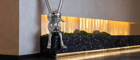 Eine metallische Hasenstatue steht auf einer schwarzen Marmorbasis, vor einer Wand mit einer Nische, in der sich eine kleine grüne Pflanze und eine spiegelnde Fläche befinden.