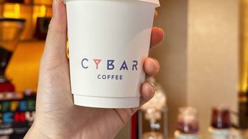 Una mano sosteniendo una taza CYBAR COFFEE con tapa blanca.
