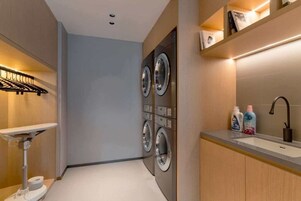 A modern laundry room with a sink, two wall-mounted washing machines, and a shelf with cleaning supplies.