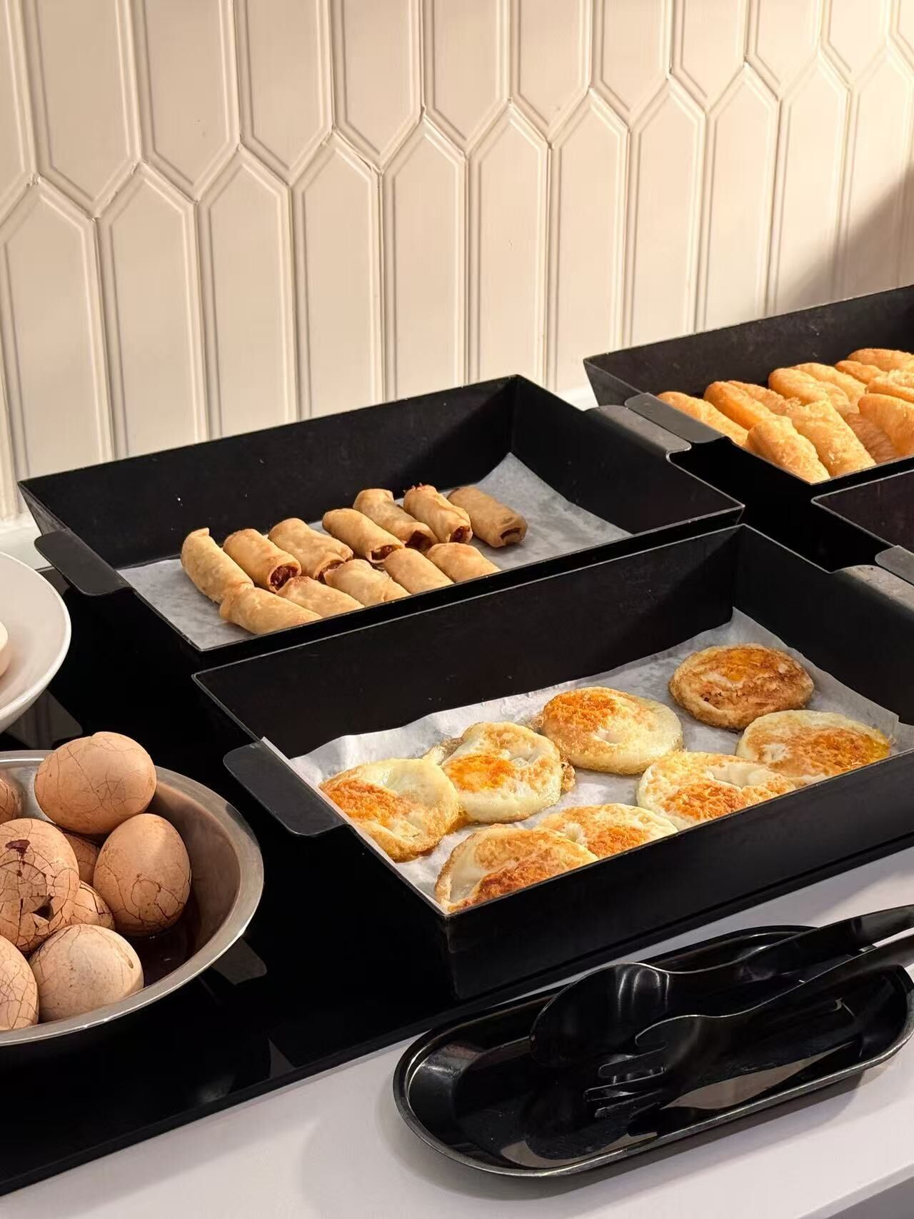 Un bufé con diversos alimentos, incluyendo rollitos primavera y pasteles, dispuestos en bandejas negras.