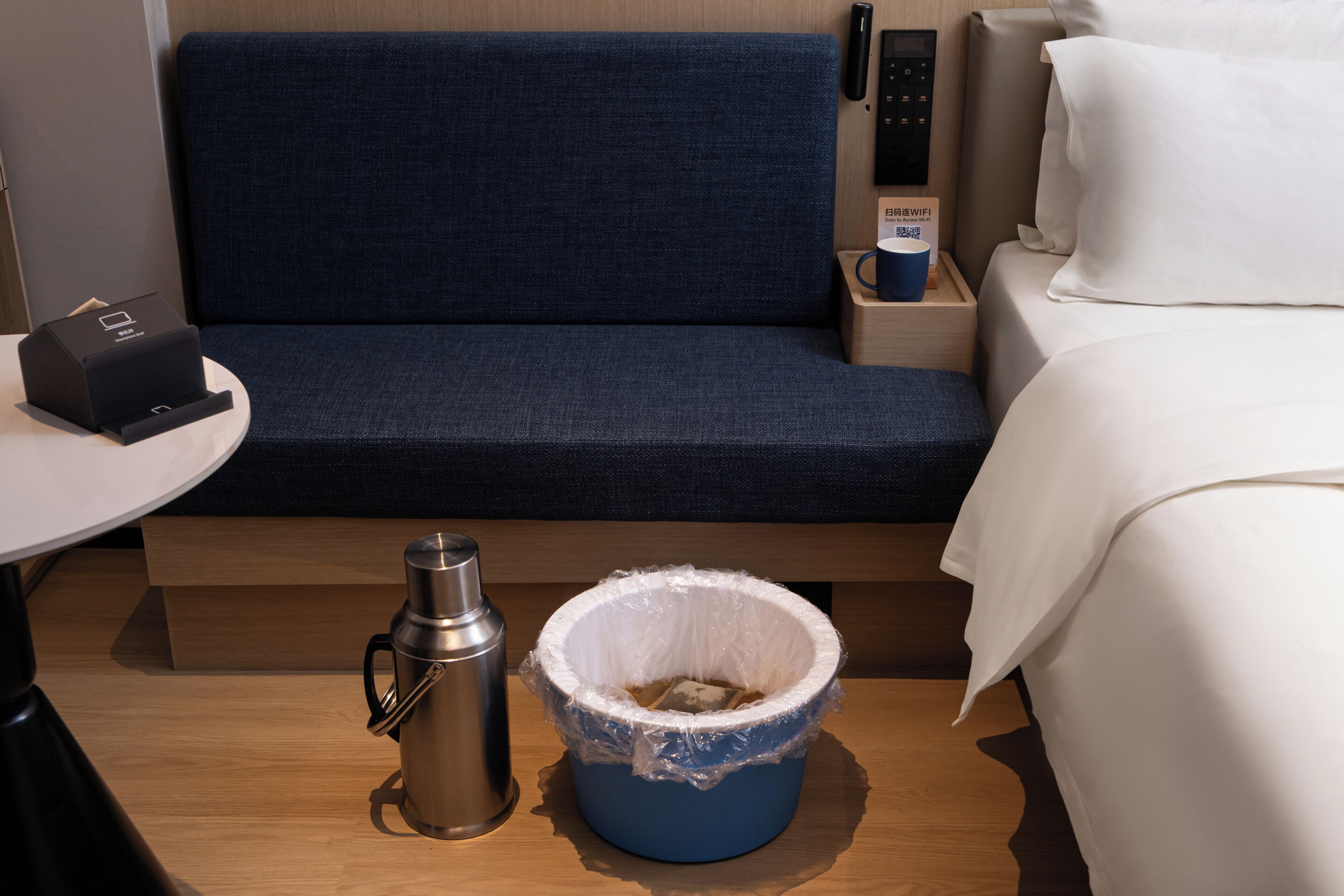 A hotel room with a bed, a small table with a coffee maker, a trash bin, and a control panel on the wall.