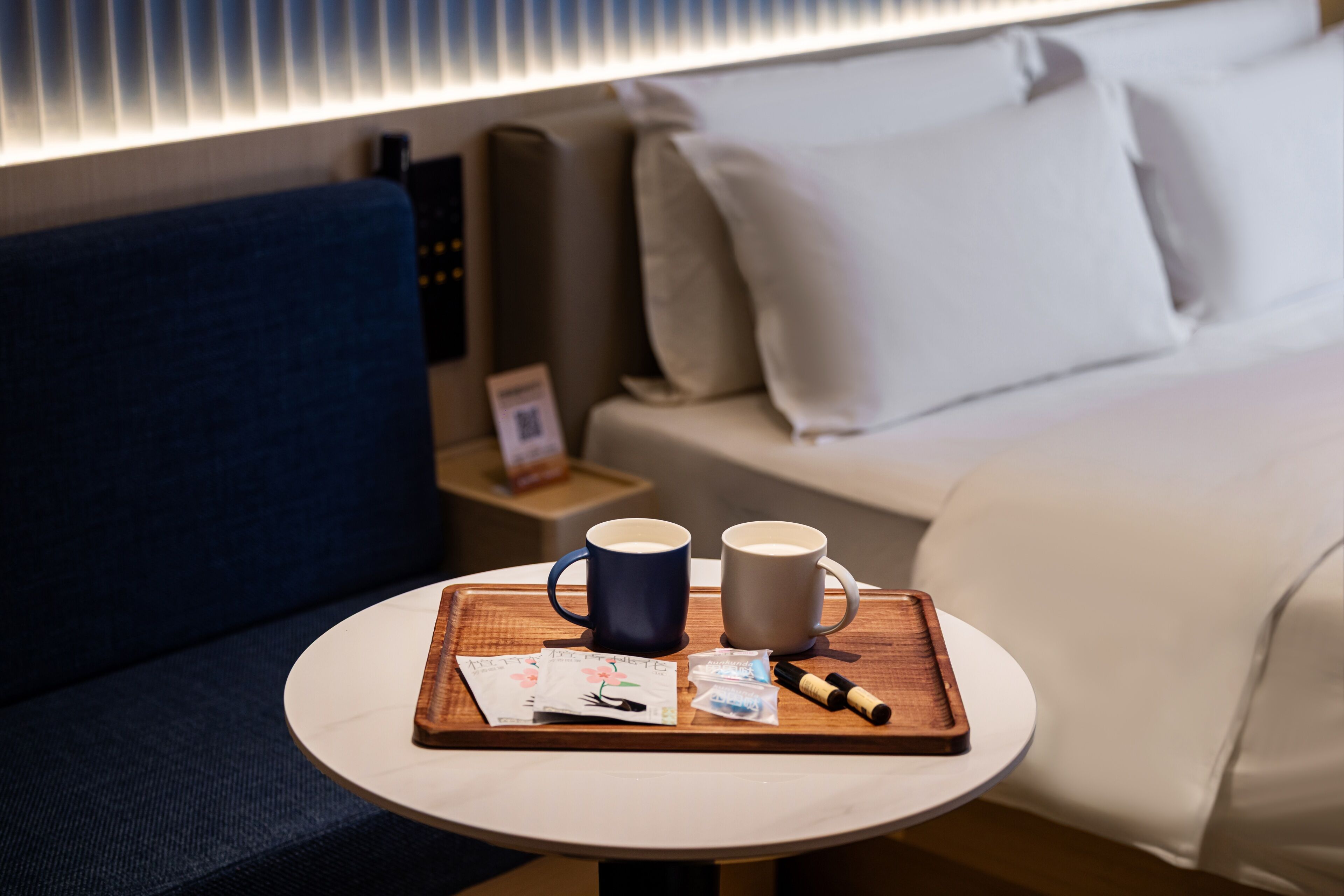 Quarto de hotel com cama, mesa de cabeceira com um tabuleiro contendo duas chávenas, caneta e algumas cartas.