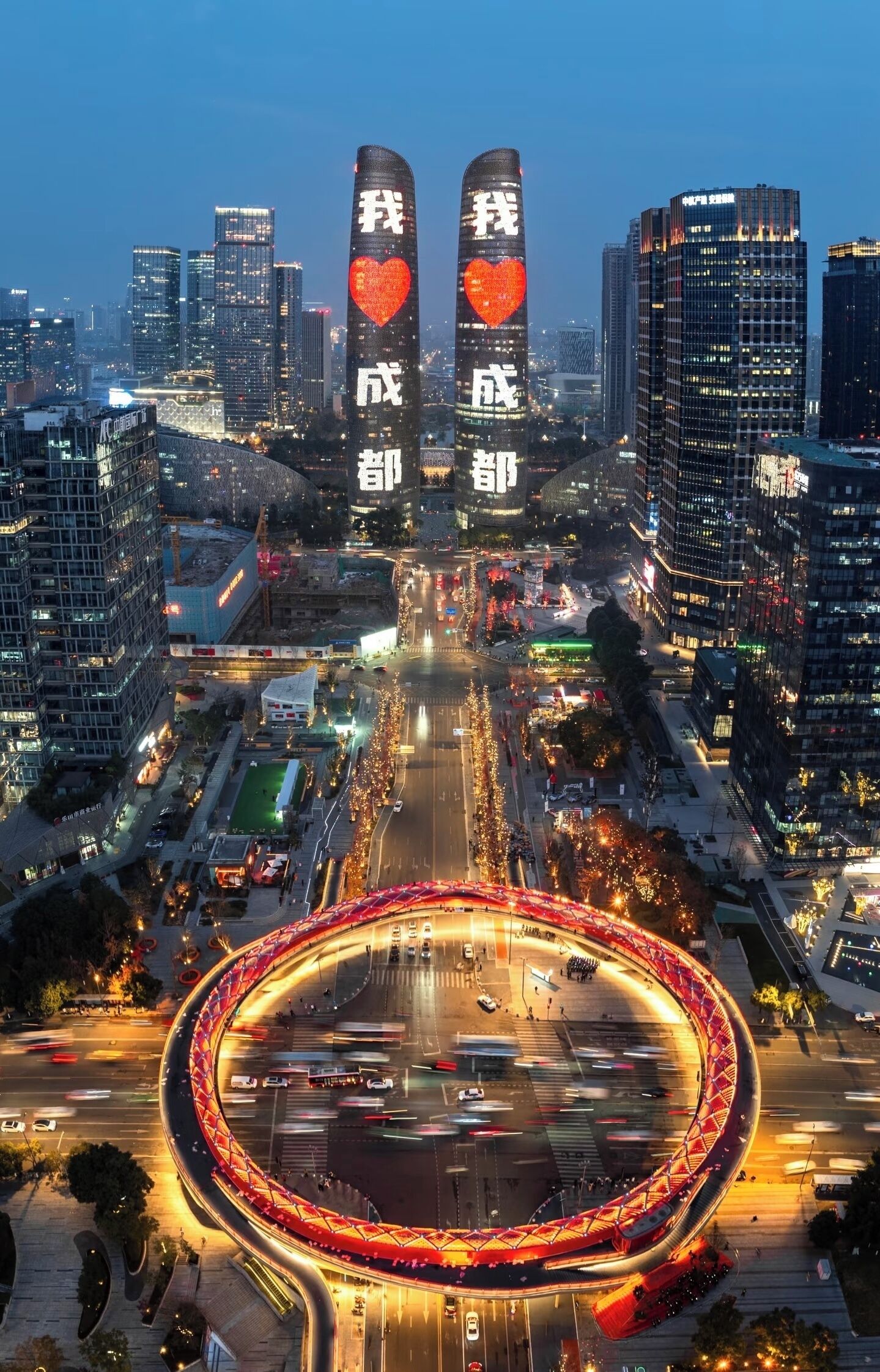 A cityscape with two prominent buildings featuring illuminated text and heart symbols, a circular road with red lighting, and surrounding modern high-rise buildings.