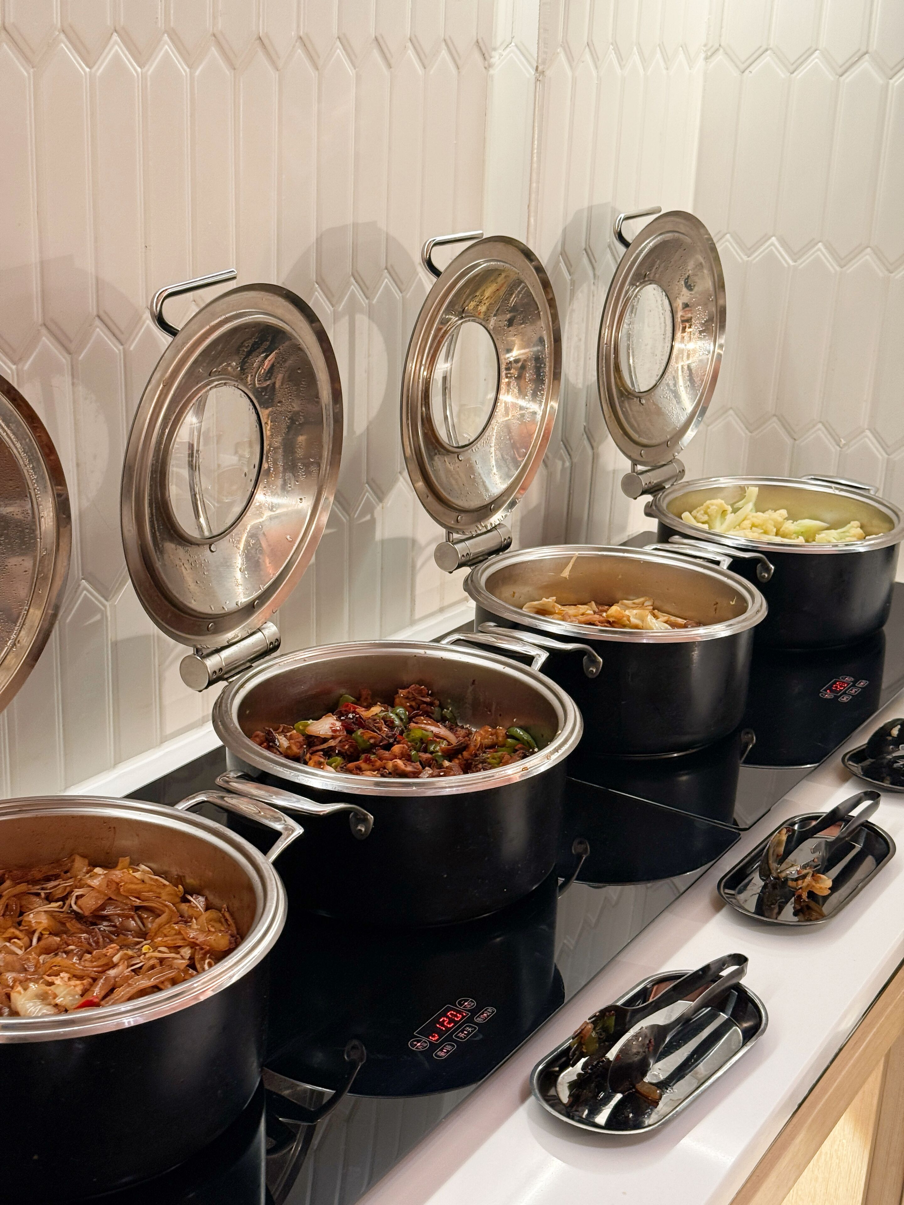 A buffet setup with multiple stainless steel pots containing various dishes, arranged on a countertop.