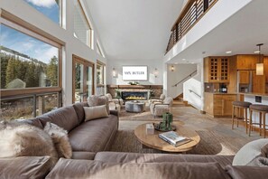 A spacious living area with a fireplace, a kitchen with wooden cabinets, and a large window overlooking a forested area. - 11 Bedroom Duplex - Vail Village (Vail)