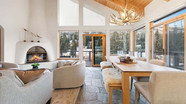 En modern inredning med en öppen spis, stengolv, en matplats och stora fönster med utsikt över skogen.