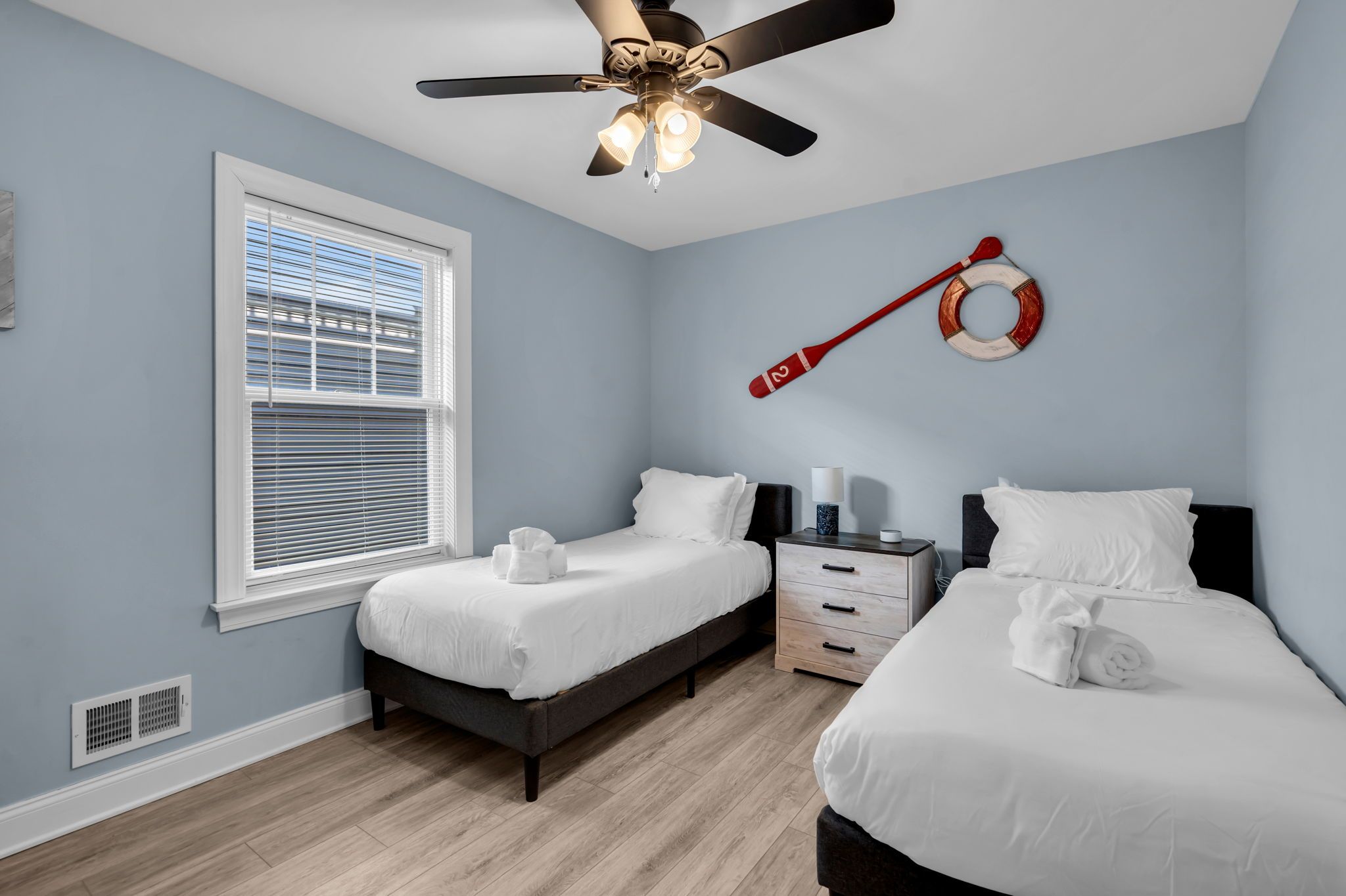 A bedroom with two beds, a ceiling fan, a window with blinds, a nightstand, and a wall-mounted life preserver.